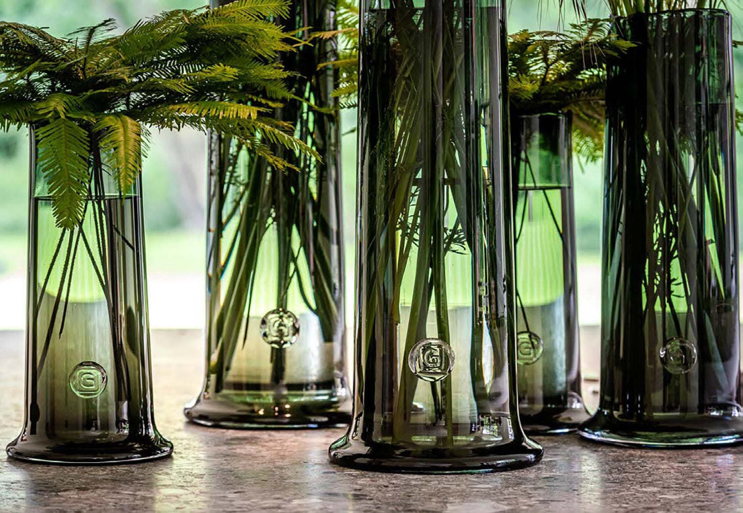 Gommaire Vase Nikky in den Größen Small, Medium und Large aus mundgeblasenem grauen Glas, gefüllt mit grünen Zweigen. Stilvolle Vasen im modernen Design.