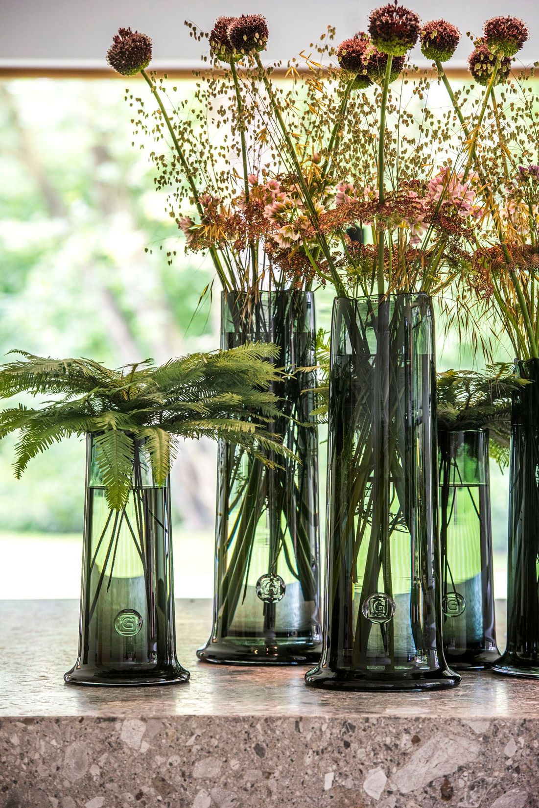 Gommaire Vase Nikky in den Größen Small, Medium und Large aus mundgeblasenem grauen Glas, gefüllt mit grünen Zweigen. Stilvolle Vasen im modernen Design.