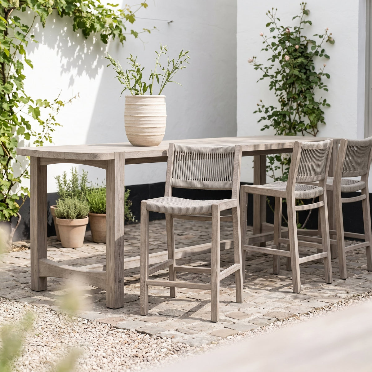 Barstuhl Mario Teak Natural Grey von Gommaire, Teak Natural Grey und Seilgeflecht, B 45 × T 56 × H 97 cm, am Bartisch Jacoba mit Vase Simon im Innenhof