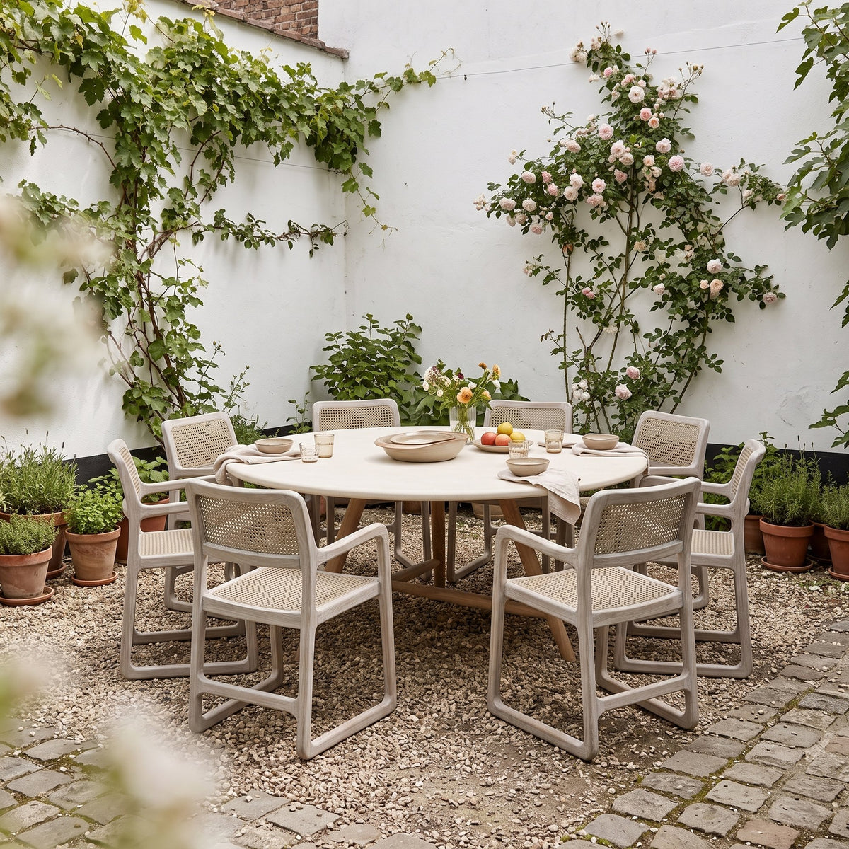 Esstisch Faye rund Beton Teak Natural Grey XL von Gommaire, Beton und Teak Natural Grey, Ø 170 × H 76 cm, Ambiente mit Armlehnstühlen Sally Open Weave und Schale Ali im Innenhof