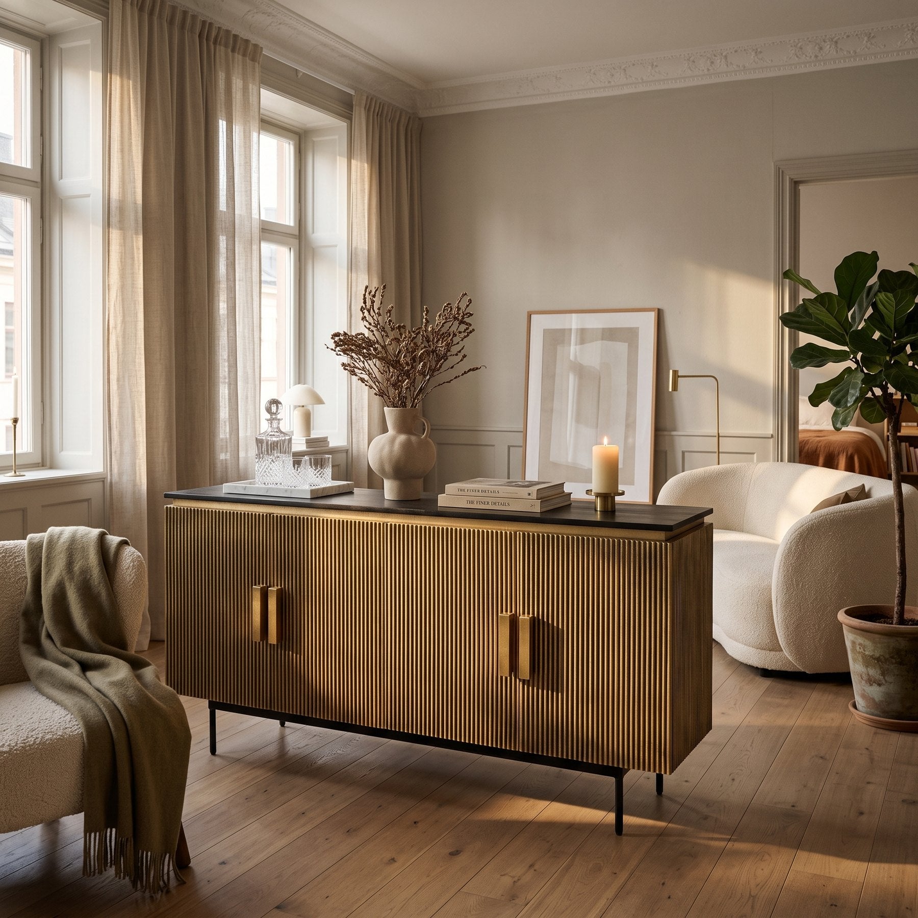 Sideboard Virat in antiker Messingoptik mit dunkler Mangoholz-Platte an der Wand eines hellen Altbau-Wohnzimmers, daneben ein Bouclé-Sofa und eine Feige
