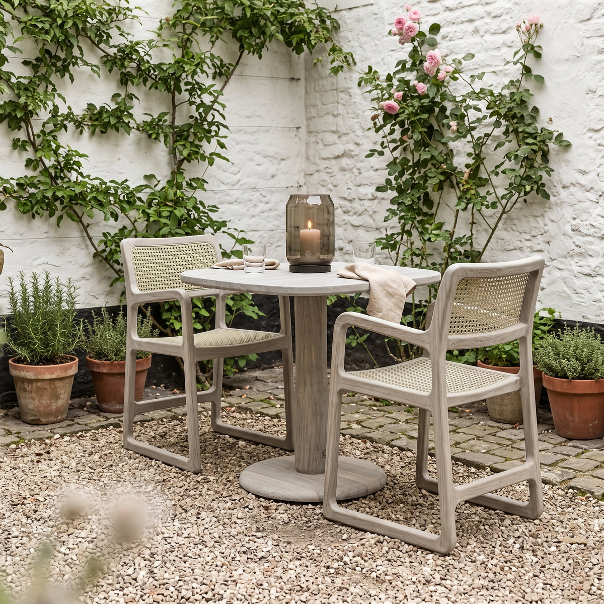 Armlehnstuhl Sally Open Weave Teak Natural Grey von Gommaire, Teak Natural Grey und PE-Rattan, B 52 × T 60,5 × H 80,5 cm, mit Bistrotisch Dita und Windlicht Daniel im Innenhof