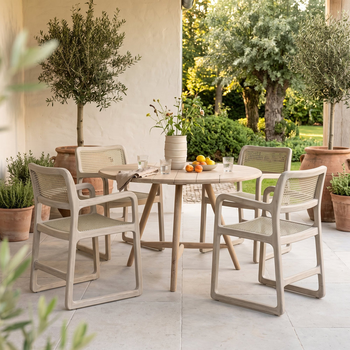 Armlehnstuhl Sally Open Weave Teak Natural Grey von Gommaire, Teak Natural Grey und PE-Rattan, B 52 × T 60,5 × H 80,5 cm, am runden Esstisch Faye mit Vase Simon auf der Terrasse