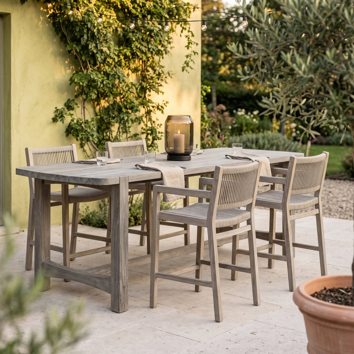 Barstuhl Mario mit Armlehnen Teak Natural Grey von Gommaire, Teak Natural Grey und Seilgeflecht, B 52 × T 61,5 × H 97 cm, am Bartisch Jacoba mit Windlicht Daniel auf der Barterrasse