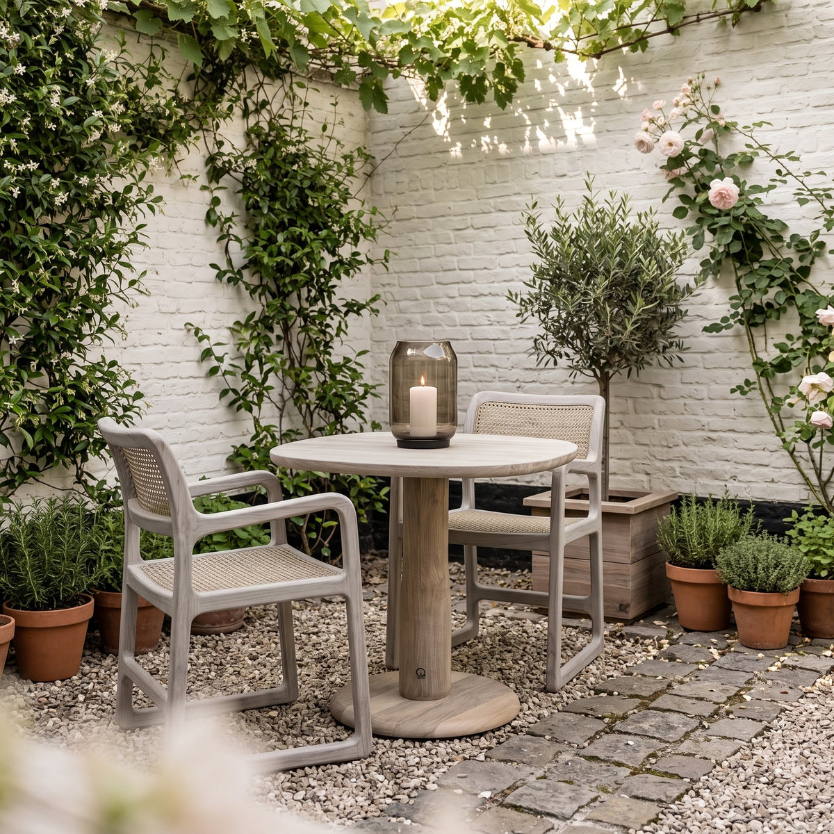 Bistrotisch Dita Squircle Teak Natural Grey von Gommaire, Teak Natural Grey, Ø 80 × H 76 cm, mit Armlehnstühlen Sally Open Weave und Windlicht Daniel im Innenhof