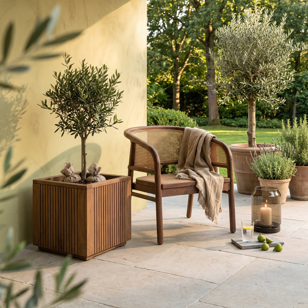 Pflanzkübel Nora Teak Emas Brown Small von Gommaire, Teak Emas Brown, 50 × 50 × H 50 cm, mit Olivenbaum neben einem Outdoor Sessel Imen und Windlicht Daniel auf der Gartenterrasse